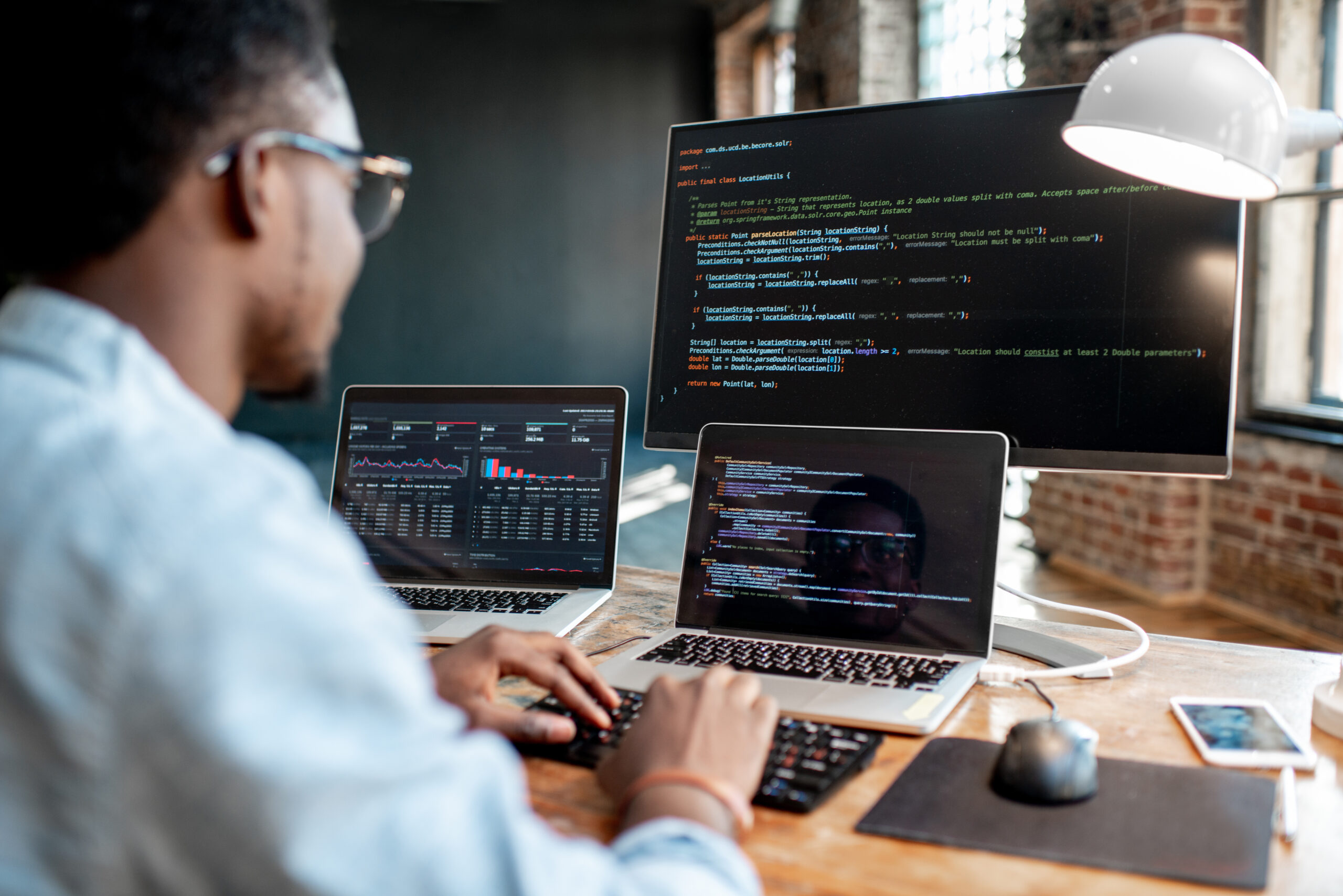 Young african american male programmer writing program code sitting at the workplace with three monitors in the office. Image focused on the screen