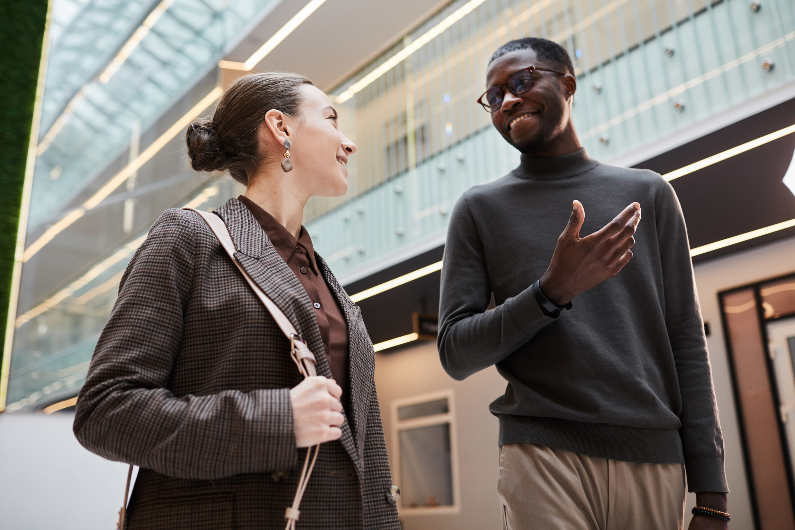 Low angle portrait of two young coworkers chatting while walking in hall at modern office building