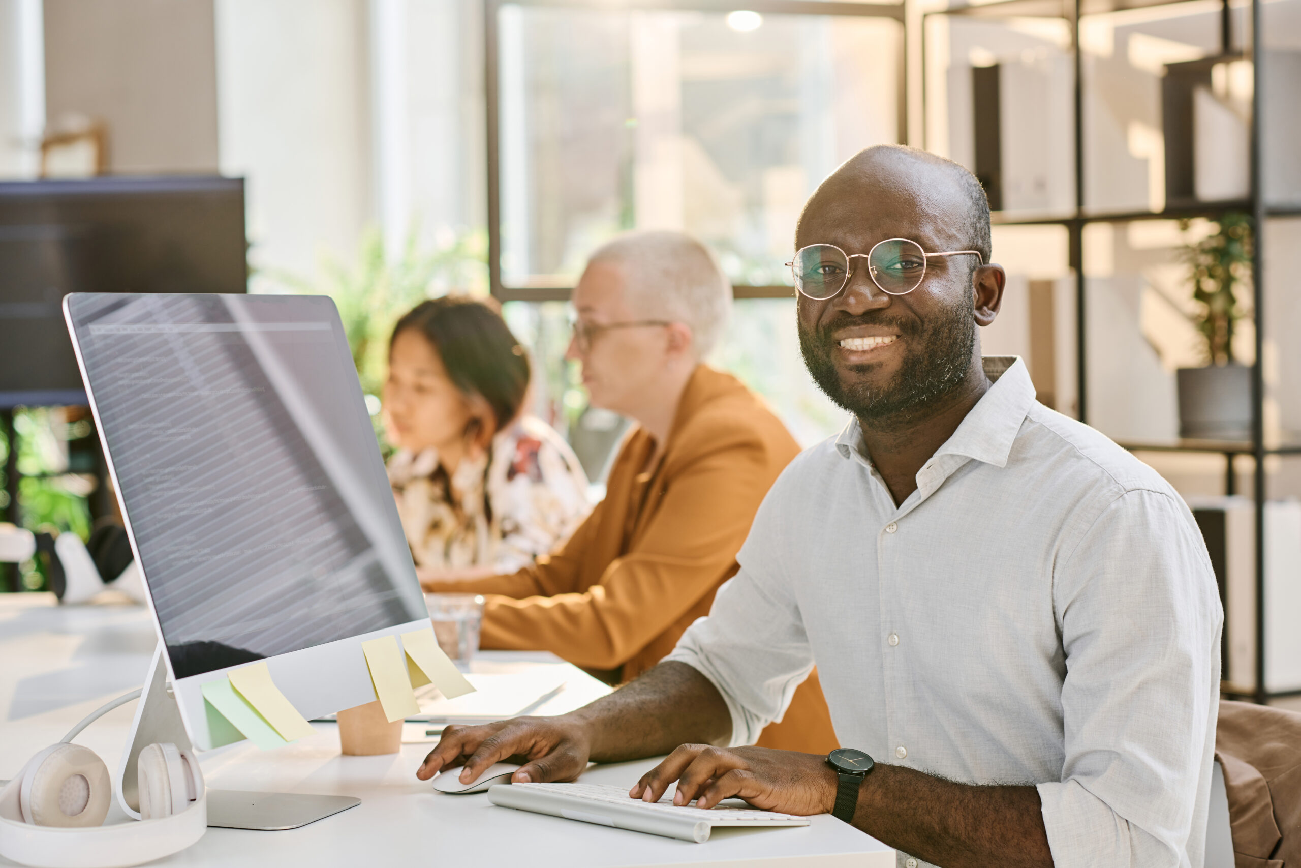 Smiling Black male programmer working on computer while sitting at desk beside colleagues in office