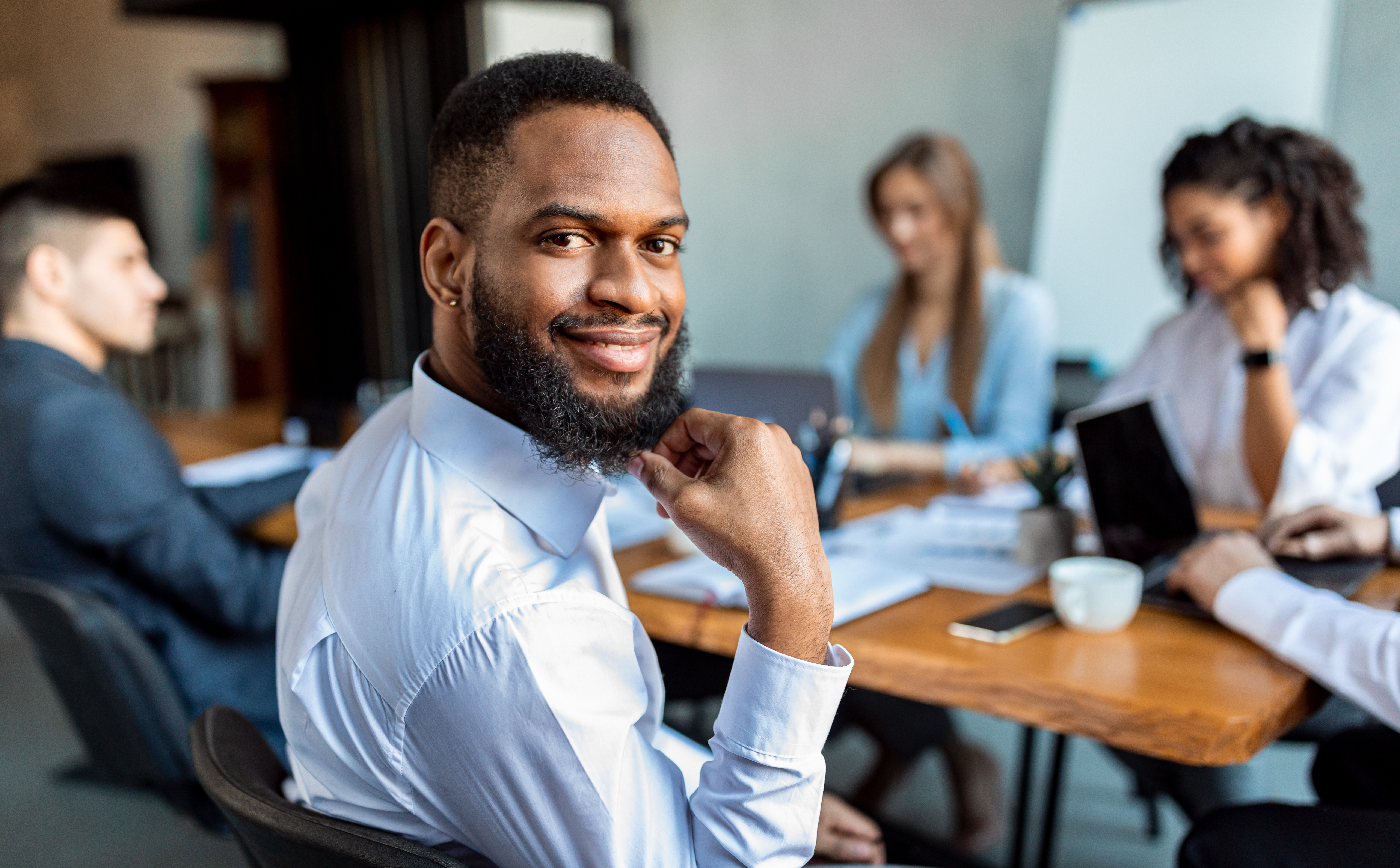 Business Meeting. African American Businessman Sitting With Colleagues Smiling To Camera During Negotiations In Modern Office.